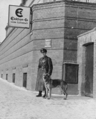 Bis 1938 hieß das Unternehmen Elektronmetall. Werkschutzmann mit Hund vor einem Eingang um 1936.